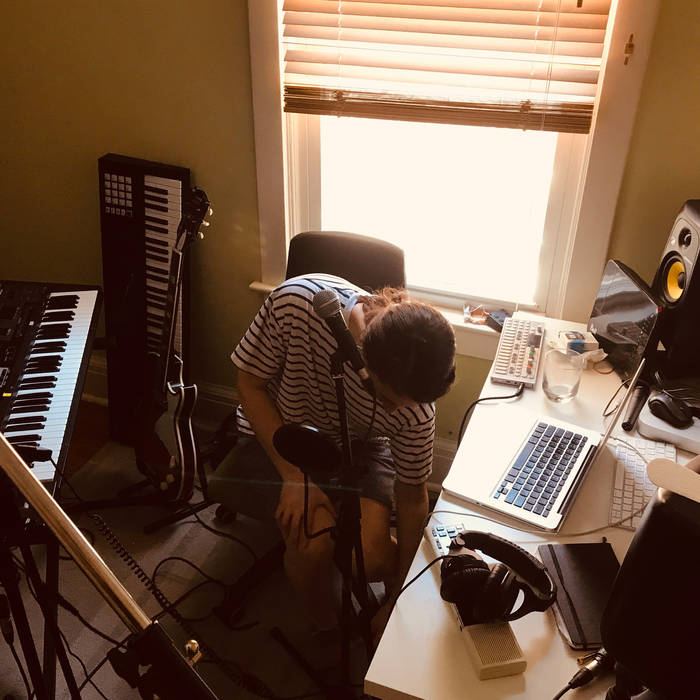 image is a photograph of the artist seated in front of a window of a home recording studio surrounded by instruments and equipment. The artist is bending at the waist, looking down as they reach for something unseen on the floor